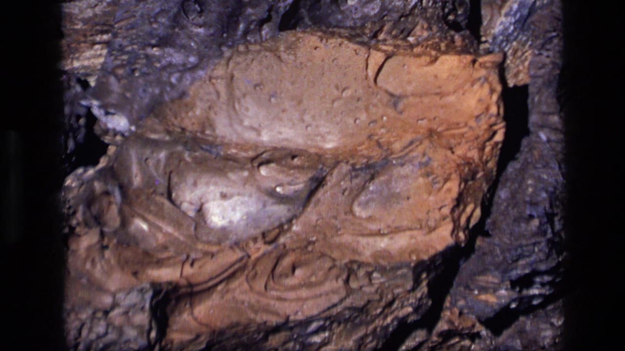 1972 Idaho 8mm frame showing a tight close-up of pigmented rock with brown, purple, and glossy mineral-like surfaces.