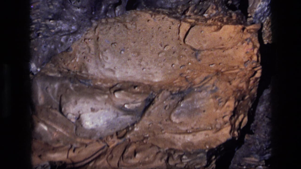 1972 Idaho 8mm frame with mottled brown rock, dark surrounding stone, small pits, and reflective mineral-like patches.