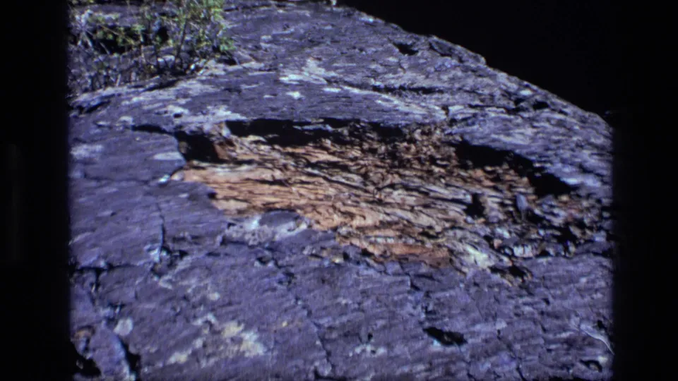 1972 Idaho 8mm frame showing the zoomed-out rock surface, with dark weathered stone, exposed layered texture, and small vegetation at the top.