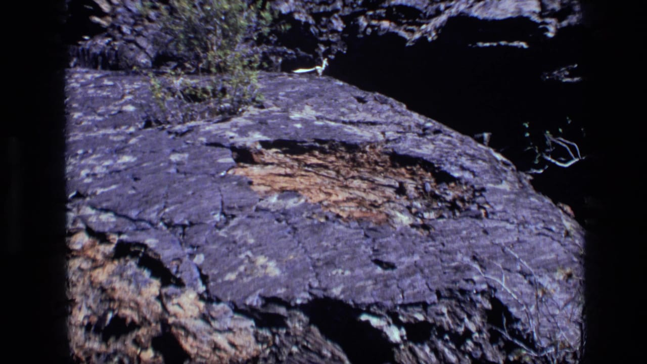 1972 Idaho 8mm frame with purple-gray layered rock, visible cracks, and a dark shadowed background around the stone.