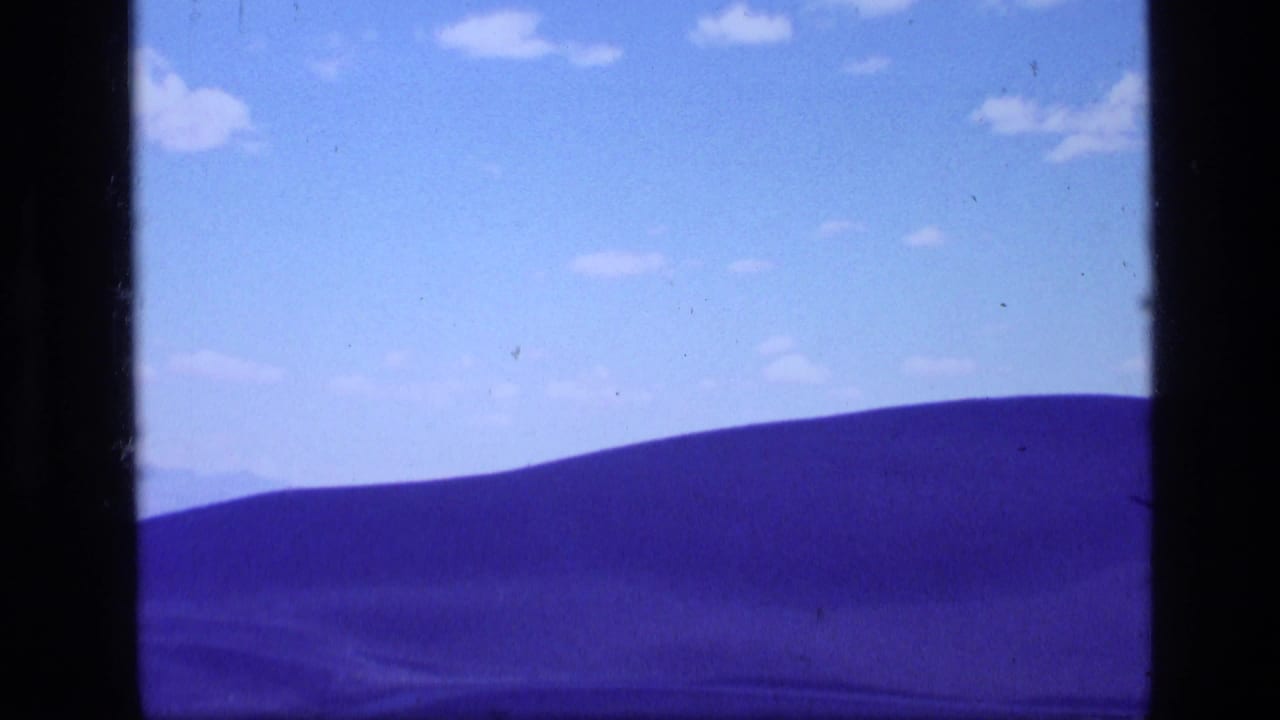1972 Idaho 8mm frame of a rolling mountain ridge beneath a bright blue sky with small clouds.