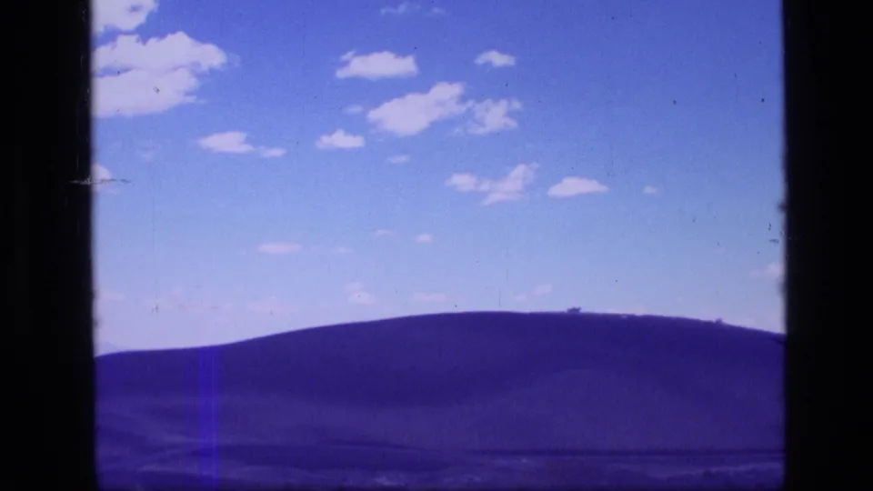 1972 Idaho 8mm frame of a rolling hillside with purple-blue color shift beneath a cloud-dotted sky.