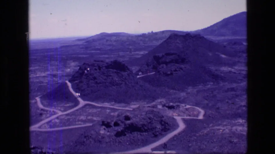 1972 Idaho 8mm frame showing arid hills, volcanic-looking rocky forms, winding dirt roads, and a hazy open sky.