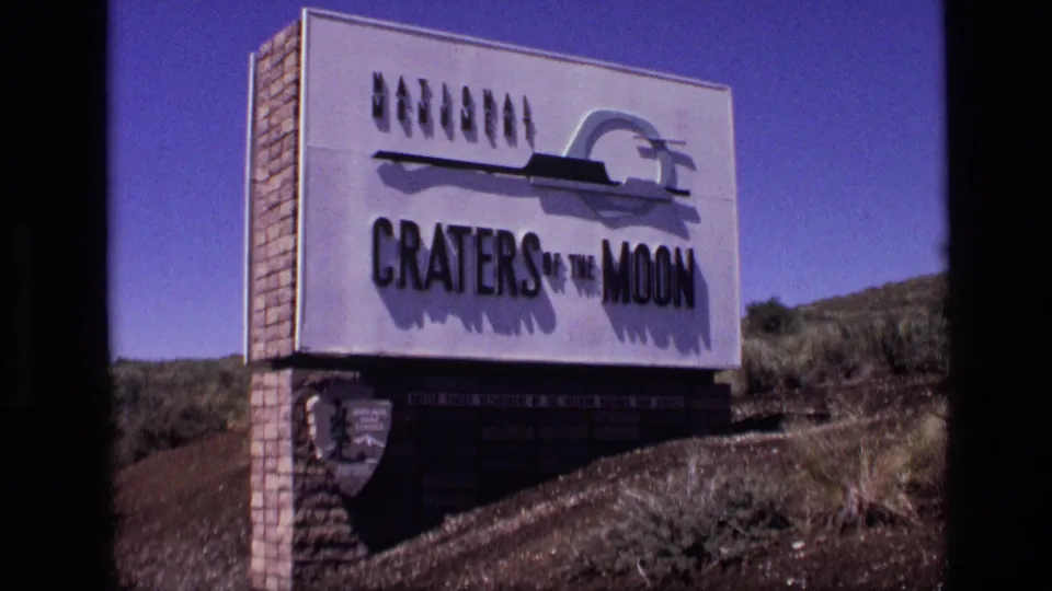Visitor-shot 8mm frame of the Craters of the Moon sign in Idaho with dry terrain, scrub, brick supports, and blue sky.