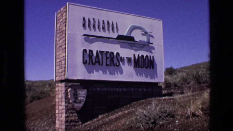 1972 8mm frame of the Craters of the Moon National Monument sign in Idaho, with brick base, NPS shield, and arid hillside.
