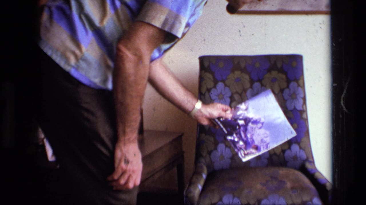 Man reaches toward a floral armchair while holding a magazine in a dim living room.