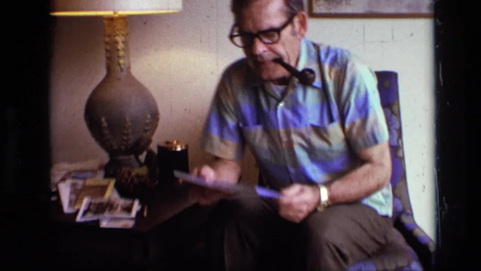 A man wearing glasses smokes a pipe and sits with a magazine near a table lamp and scattered photographs in a 1973 home interior.