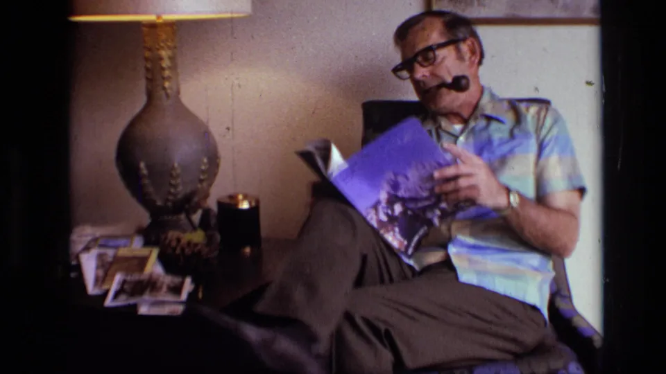 The man reclines in a floral armchair, pipe in mouth, reading a magazine beside a lamp and small pile of photographs.
