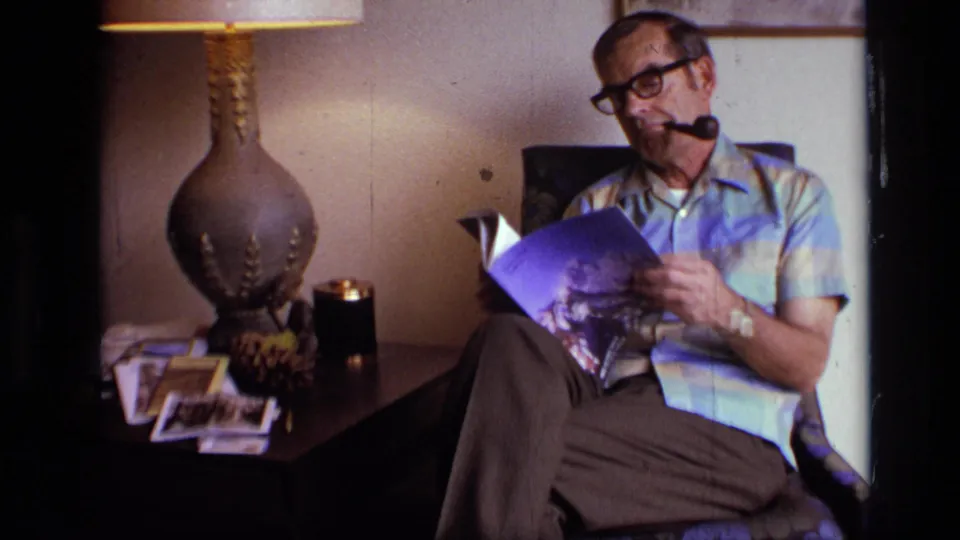 A seated man in glasses reads a magazine with a pipe in his mouth, framed by a lamp, side table and floral armchair in a private interior.