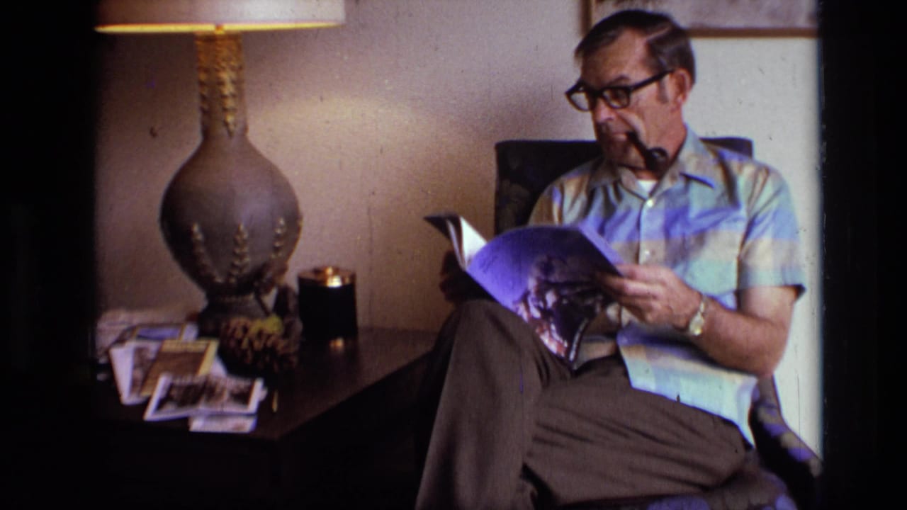 Older man with glasses and a pipe reads a magazine beside a lit table lamp.