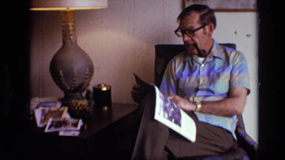 1973 8mm frame of an older man with glasses and a pipe shifting a magazine page in a private living room.