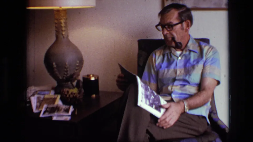 1973 home-movie frame with an older man seated in an armchair, pipe in mouth, magazine open on his lap near a table lamp.