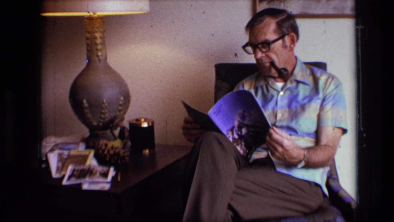 A man in a striped short-sleeve shirt reads indoors beside a lamp, side table photographs, and a candle-like object in 1973 home-movie footage.