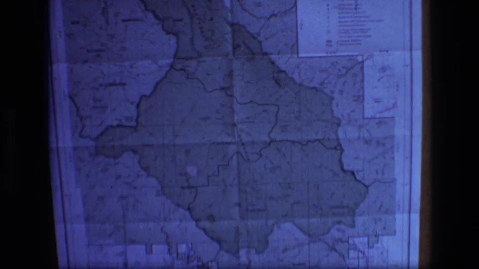 Blue-toned 8mm view of a projected Montana wilderness map, with map labels, boundary lines, and dark projector framing visible.