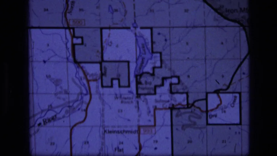 1973 Montana wilderness home-movie frame showing a purple-tinted topographic map with roads, borders, grid markings, and labels including Kleinschmidt Flat.