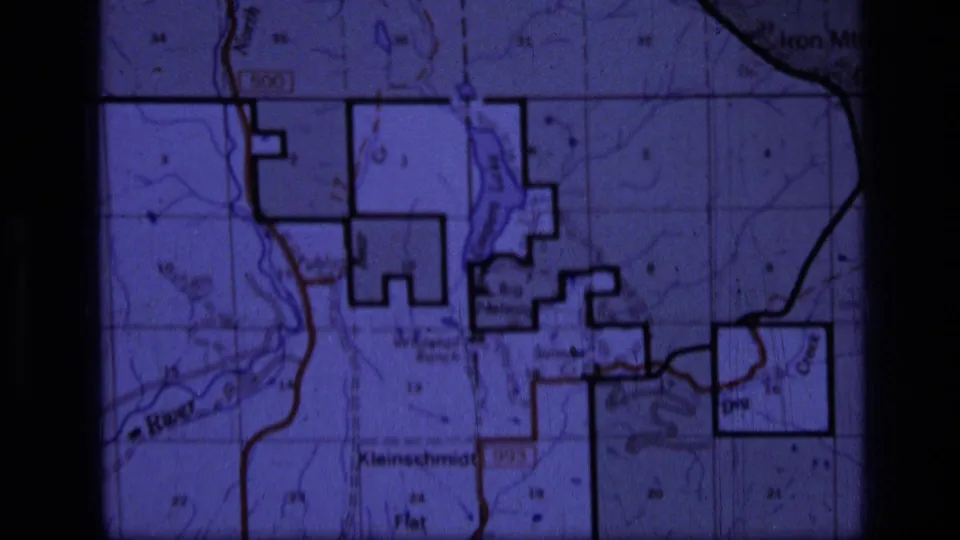 1973 Montana excursion footage holds on the topographic map, with dark boundary lines, roads, faint terrain detail, and place names visible through film grain.