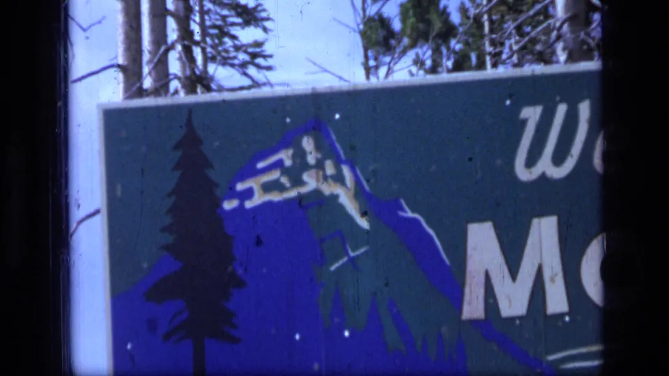 1973 Montana wilderness travel frame showing a green sign with a blue mountain and pine tree illustration, partial welcome text, and wintery speckled surface marks.