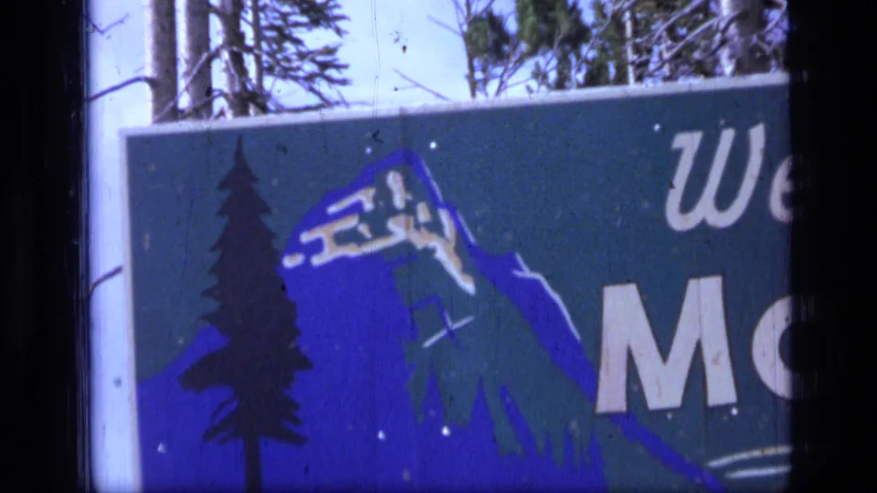 1973 Montana home-movie frame shows the sign cropped tighter, emphasizing the blue mountain artwork, dark pine tree, partial welcome lettering, and outdoor trees behind it.