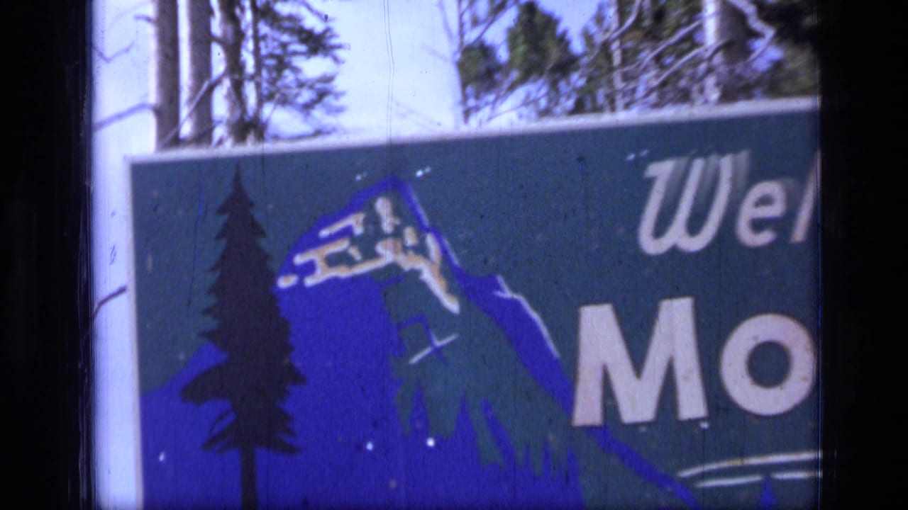 1973 8mm frame showing a cropped Montana welcome sign with blue mountain and pine graphics beside tall trees.
