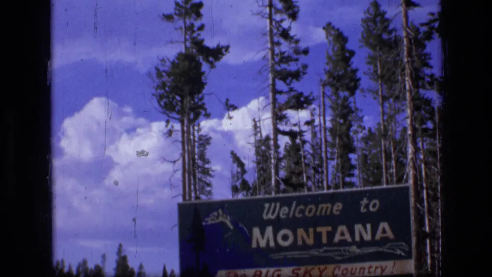 1973 8mm frame showing the Montana welcome sign below tall trees and large white clouds.