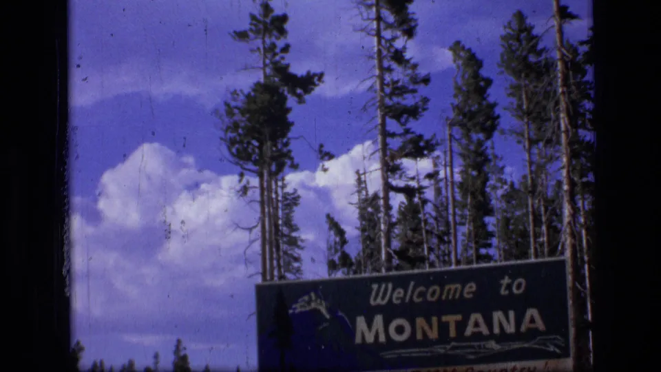 1973 8mm frame showing a wide roadside view of the Montana welcome sign with evergreens and clouds.