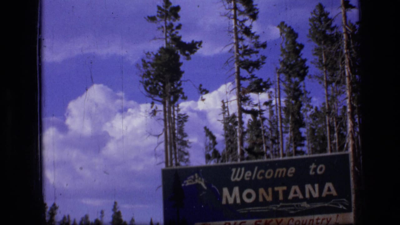 1973 8mm Montana road-trip frame showing a Welcome to Montana sign with pine trees, blue sky, and white clouds.