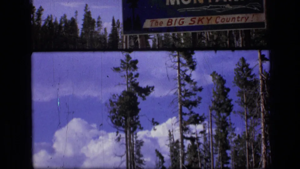 1973 Montana home-movie frame with pines, clouds, and cropped portions of the Big Sky Country welcome sign.