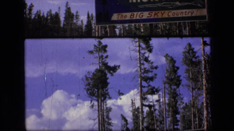 1973 Montana 8mm frame showing a Big Sky Country roadside sign over pine trees, blue sky, and white clouds.
