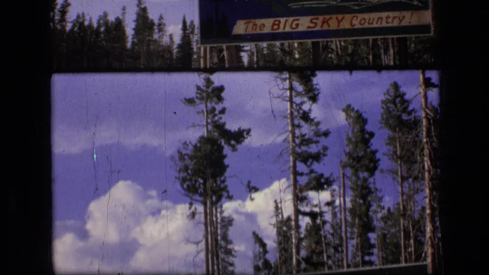 1973 Montana home-movie frame with tall evergreen trunks, cumulus clouds, and the upper edge of a Big Sky Country sign.