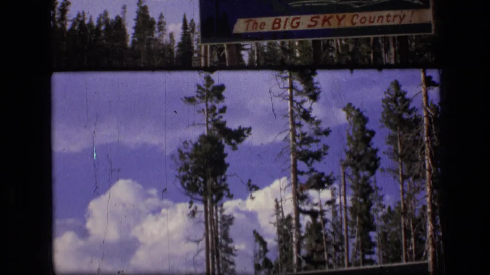 1973 Montana 8mm frame showing a moving view of evergreen forest, cloud-filled sky, and a cropped Big Sky Country sign.