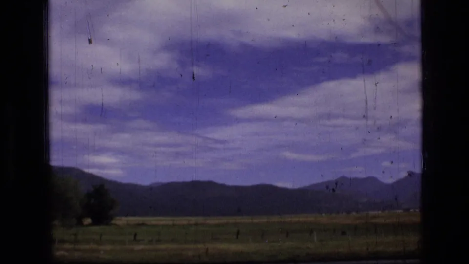 Montana wilderness landscape with a meadow, fence posts, dark hills, distant mountains, and broad clouds on 8mm film.