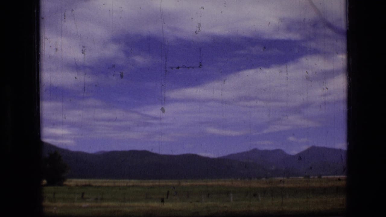 Rural field and distant mountains seen through a dark window frame under a clouded sky.