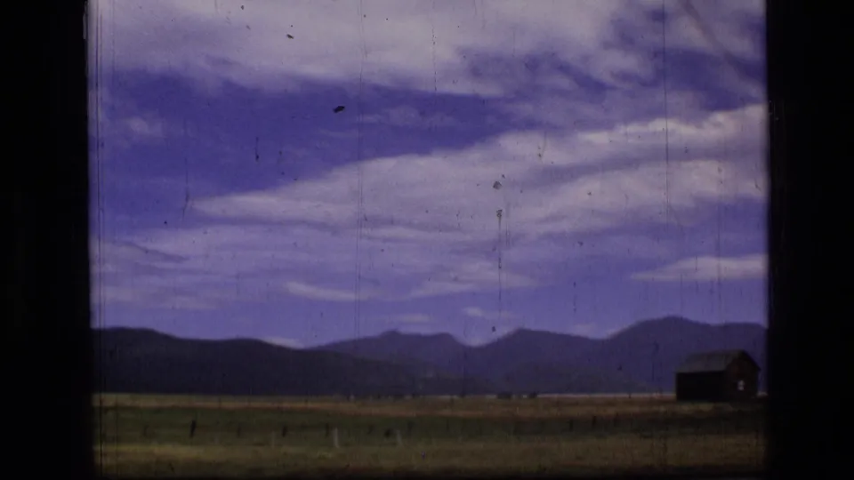 1973 Montana wilderness view with a broad field, low mountain ridge, purple-blue clouded sky, and small cabin near the right edge.