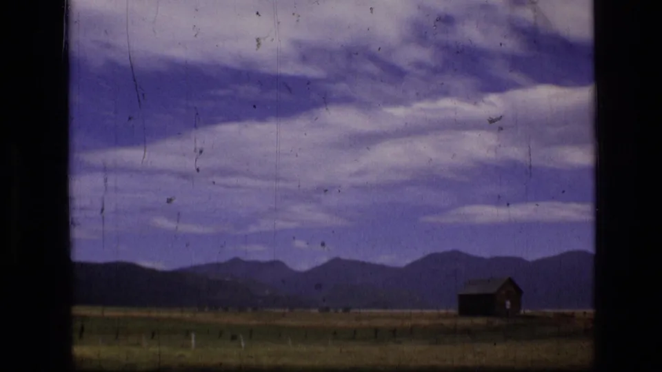 1973 8mm view of a small wooden cabin across an open Montana field, with mountains and streaked clouds in the background.