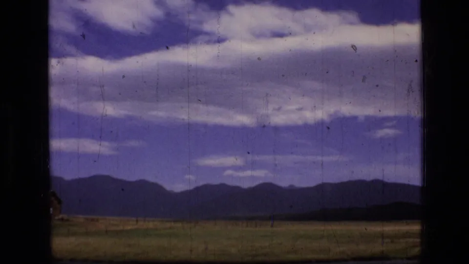 1973 Montana landscape frame with field, low mountains, layered clouds, and a dark vertical window or film-frame obstruction.