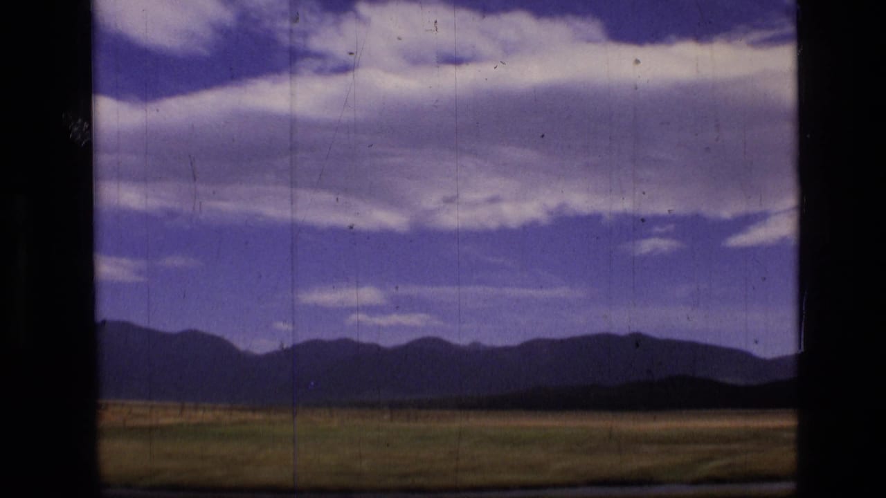 Window-framed meadow with low hills, distant mountains, blue sky, and white clouds.