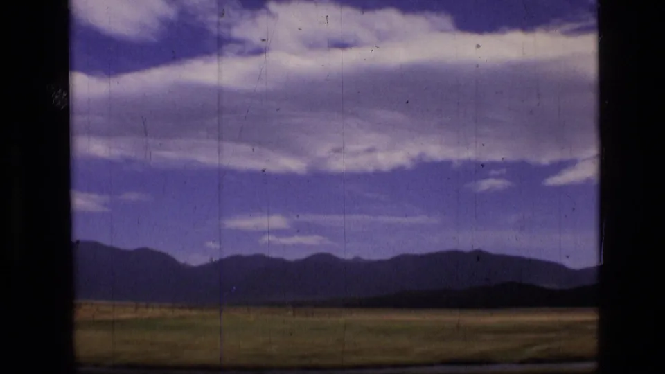 1973 8mm view of Scapegoat Wilderness, Montana, showing meadow, dark hills, distant mountains, and layered clouds through a window frame.
