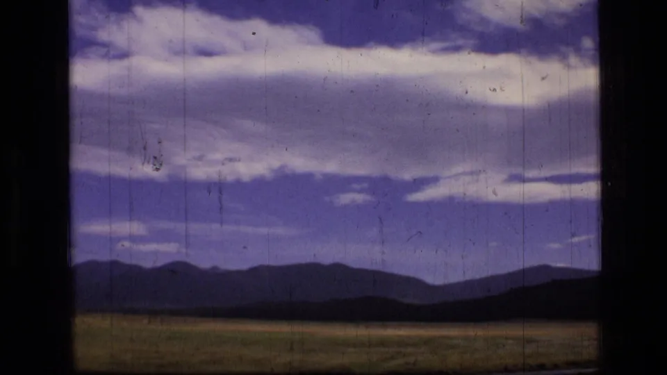 Scapegoat Wilderness, Montana, appears beyond a meadow in a 1973 8mm frame with blue sky, white clouds, and dark window borders.