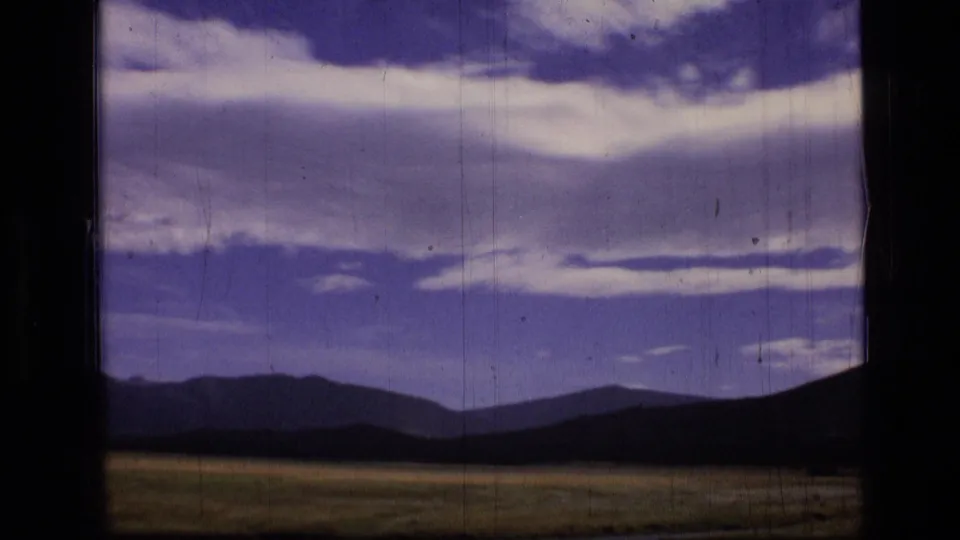 1973 Montana wilderness footage shows a meadow, shadowed hills, distant mountains, and a wide clouded sky through a window-framed view.