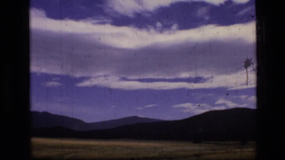 A 1973 8mm landscape frame from Scapegoat Wilderness, Montana, shows meadow, dark ridge, distant mountains, and layered clouds with film wear.