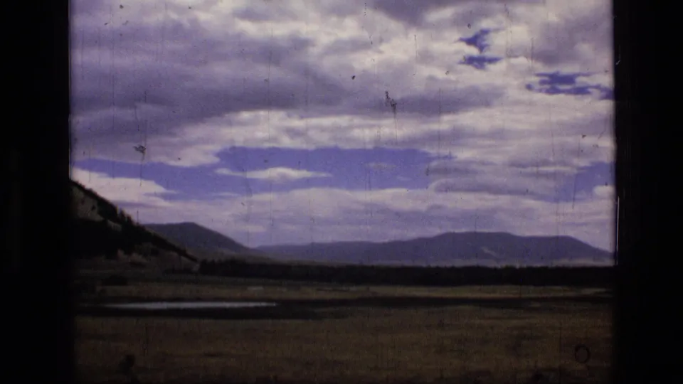 1973 8mm Montana landscape frame with open valley meadow, left hillside, wet ground, and distant mountains.