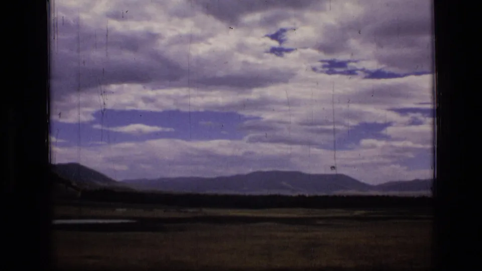 1973 8mm Montana wilderness view with dark foreground meadow, distant ridges, and dramatic cloud cover.