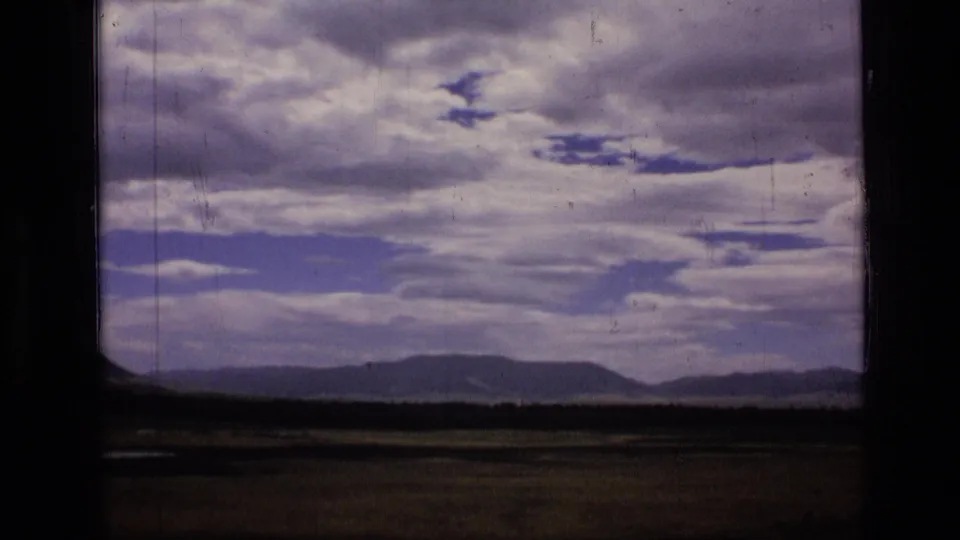 1973 8mm view of Scapegoat Wilderness, Montana, with low hills under layered clouds and a dark frame edge at left.
