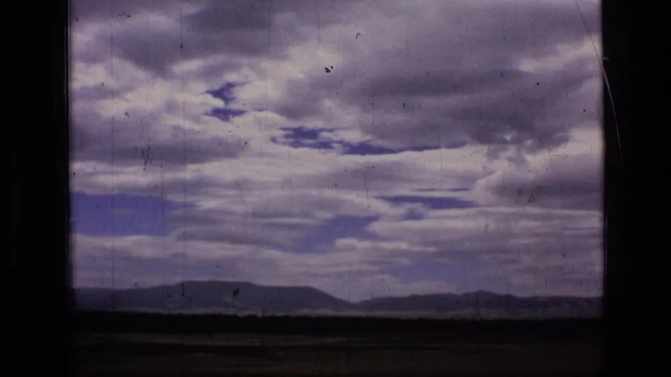 1973 8mm Montana wilderness frame showing a distant ridge, open horizon, and heavy cloud cover with blue gaps.