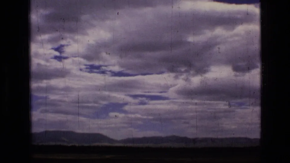 1973 8mm view of Scapegoat Wilderness with a low mountain horizon, dark open land, and textured clouds across the sky.