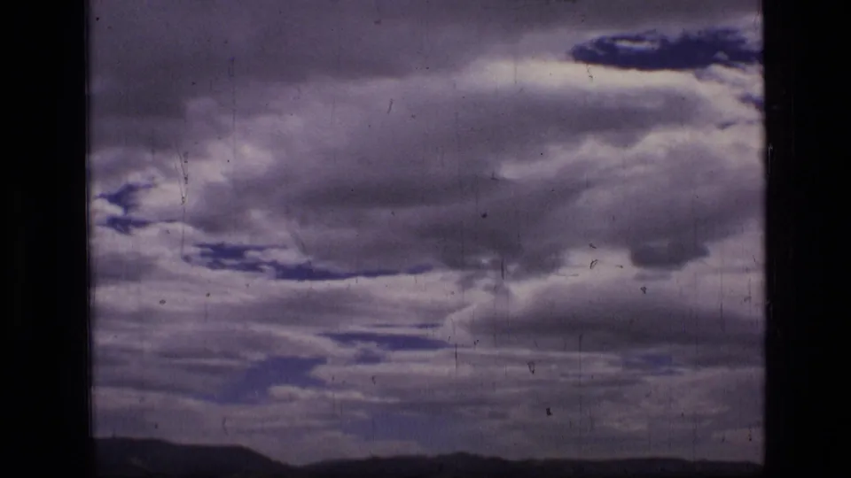 1973 8mm Montana wilderness frame with distant hills, layered clouds, blue openings, and visible film wear near the end of the pan.