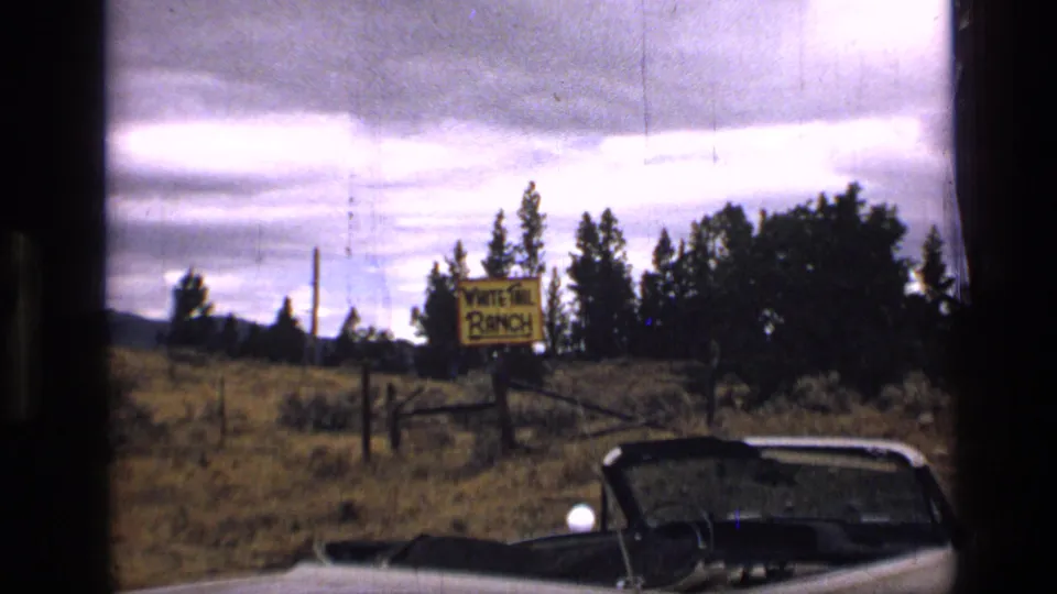 A convertible appears near a White Tail Ranch sign in a grassy Montana field, with trees and distant hills under a cloudy sky.