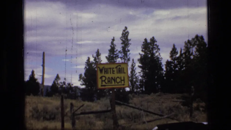 1973 8mm Montana frame with the White Tail Ranch sign more centered beside a rustic fence and evergreen trees under overcast sky.