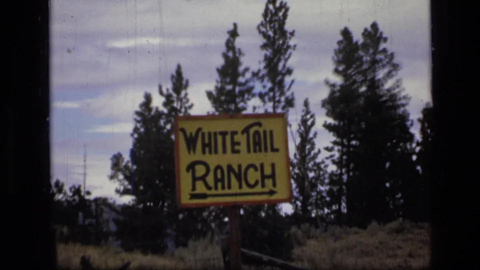 Closer 1973 8mm Montana view of the hand-painted White Tail Ranch sign, with evergreen trees, fence rails, grass, and cloudy sky visible.