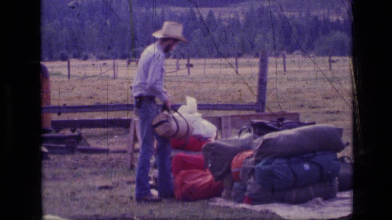 Man in a cowboy hat stands beside camping bags and sleeping rolls in a fenced field.